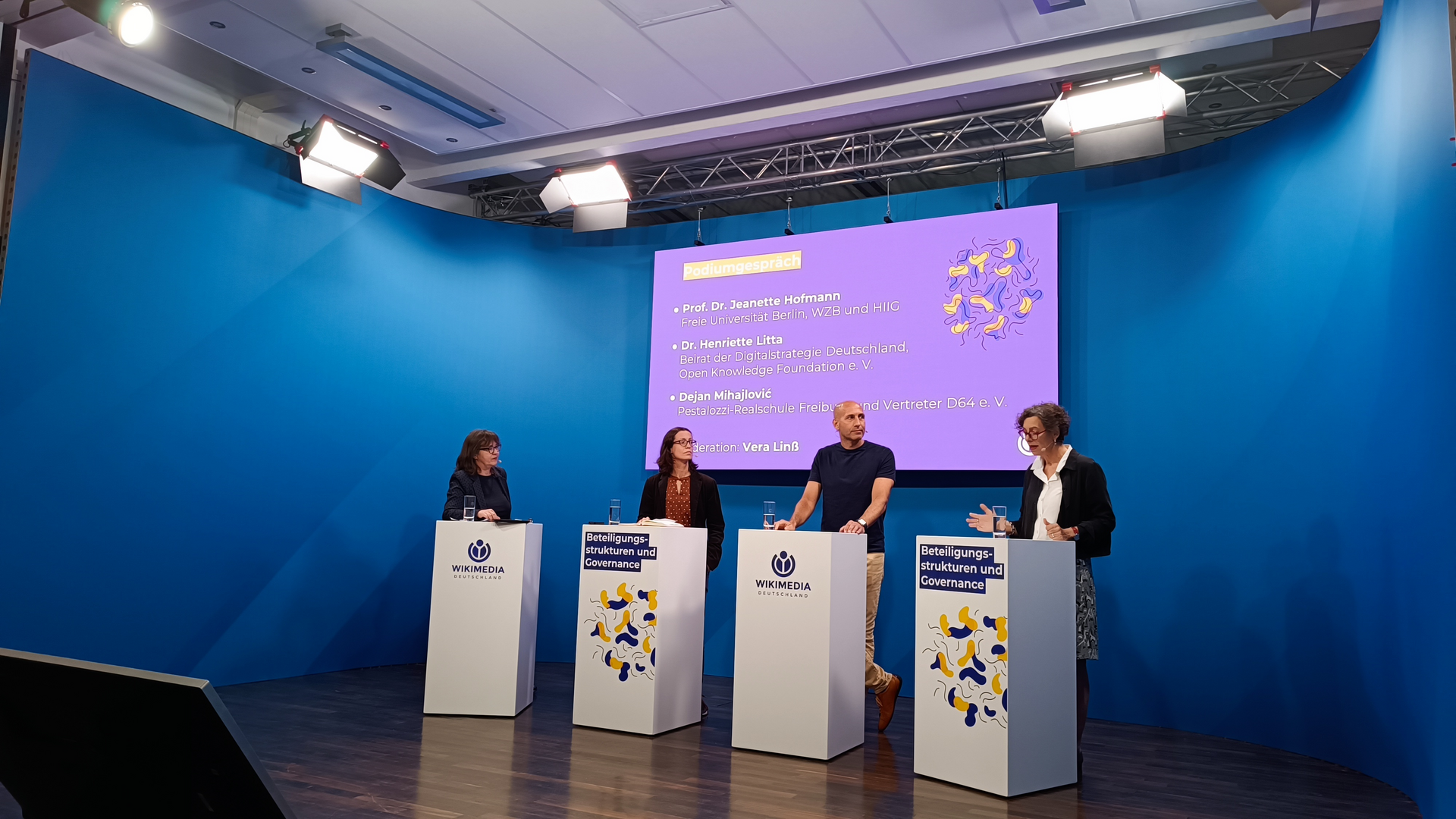 Teilnehmende der Podiumsdiskussion stehen an ihren Pulten und diskutieren.